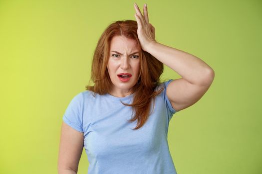 Ouch you kick my head. Frustrated concerned upset middle-aged redhead woman look shocked distressed touch temple complain kid shoot ball her face stand disappointed green background
