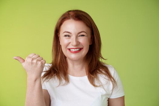 Come visit our store. Cheerful pleasant friendly charming redhead middle-aged woman entrepreneur inviting check-out promo smiling happily sincere kind grin pointing left thumb stand green background