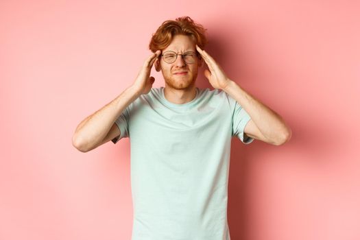 Portrait of redhead man in crooked glasses touching head and feeling dizzy or nauseous, having hangover or headache, standing over pink background