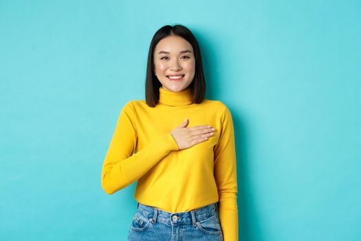 Image of proud smiling asian woman holding hand on heart, showing respect to national anthem, standing over blue background