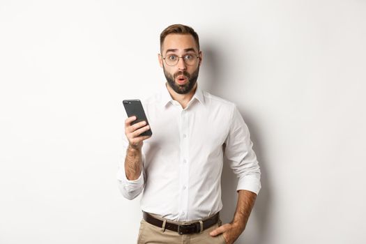 Surprised handsome man reading interesting info online, holding smartphone, standing over white background