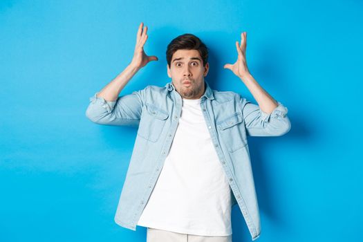 Distressed guy showing mind blowing gesture, looking frustrated and anxious, standing against blue background