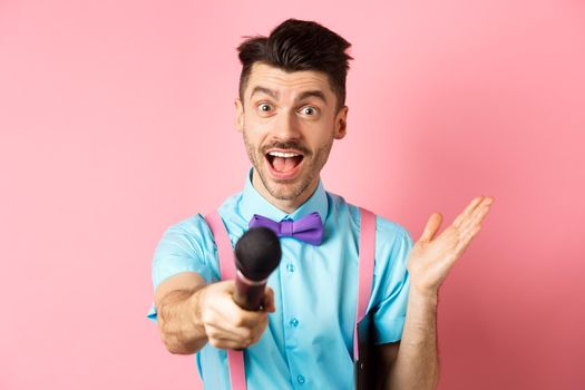 Cheerful young man, show host in bow-tie giving you microphone and smiling, entertain people on festive celebration events, performing at holidays, standing on pink background