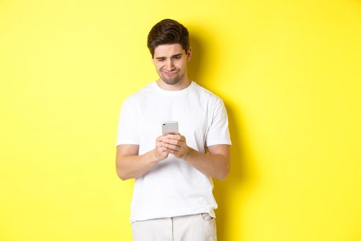 Guy looking displeased at smartphone screen, reading strange message on phone, standing in white t-shirt against yellow background