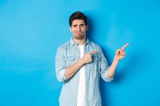Portrait of skeptical adult guy pointing fingers right and smirking, exress disappointment and doubt, standing against blue background