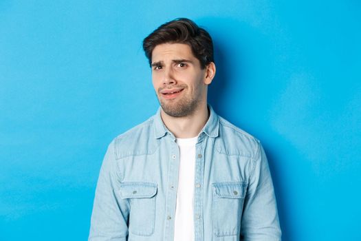 Confused and uncomfortable man looking at something strange or creepy, cringe from bad advertisement, standing over blue background