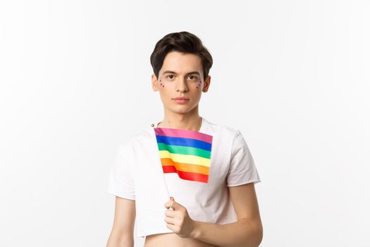 Pride and lgbtq concept. Waist up shot of attractive anrogynous man holding rainbow flag, having glitter on face and looking at camera, standing over white background