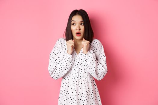 Scared and timid asian girl trembling from fear, staring left and gasping, standing in dress over pink background