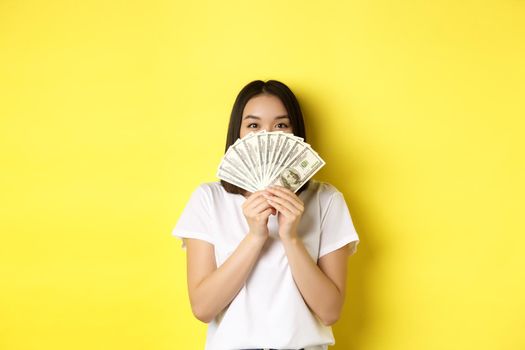 Cute asian woman hiding face behind money, peeking at camera satisfied, earn cash, standing over yellow background