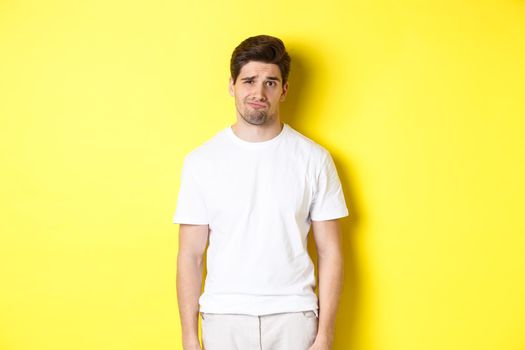 Disappointed guy looking upset, sulking and frowning, standing displeased against yellow background