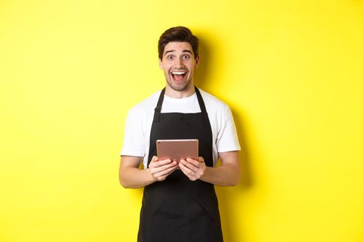 Happy salesman in black apron, holding digital tablet and looking surprised, standing against yellow background