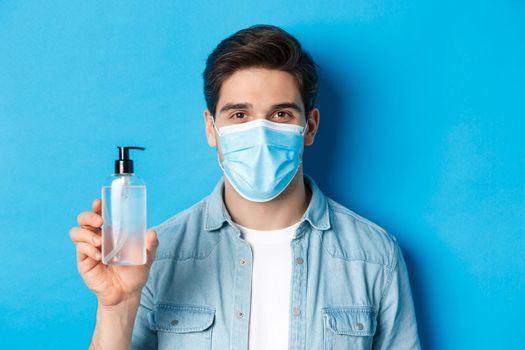 Concept of coronavirus, pandemic and preventive measures. Close-up of young man in medical mask advice to use hand sanitizer, showing antiseptic bottle, standing over blue background