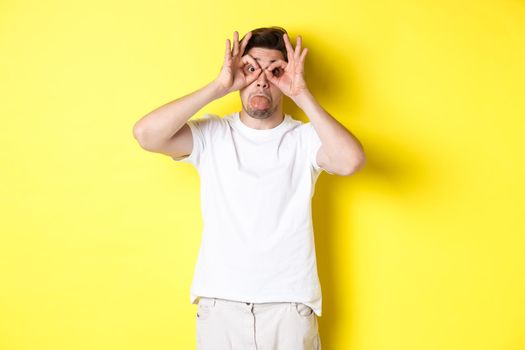 Young man making funny faces and showing tongue, fool around, standing in white t-shirt against yellow background