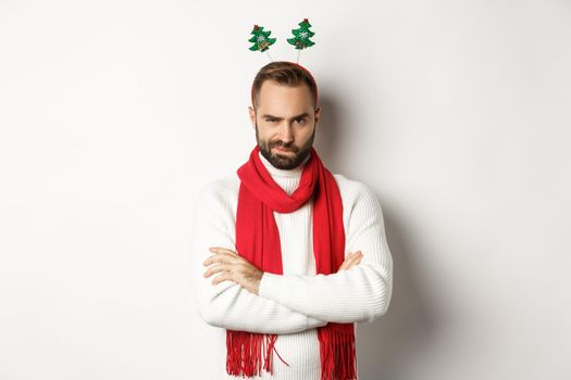 Christmas party and celebration concept. Suspicious young bearded man looking doubtful, wearing funny accessory hat, standing against white background