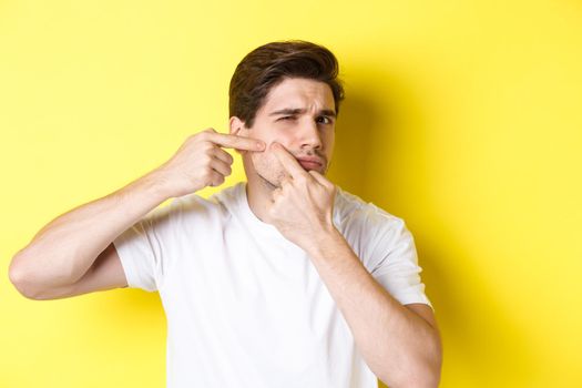 Young man pop a pimple on cheek, standing over yellow background. Concept of skin care and acne