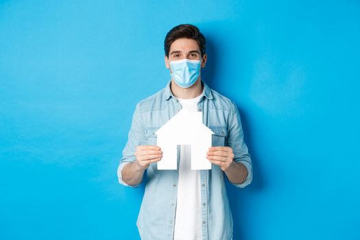 Concept of coronavirus, quarantine and social distancing. Young man searching apartment, showing house paper model, wearing medical mask, renting or buying propery, blue background