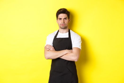 Suspicious barista squinting, cross arms on chest and looking at something with disbelief, standing over yellow background