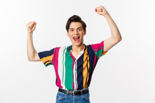 Image of happy young man triumphing of winning, celebrating victory, raising hands up in rejoice and shouting yes, standing over white background