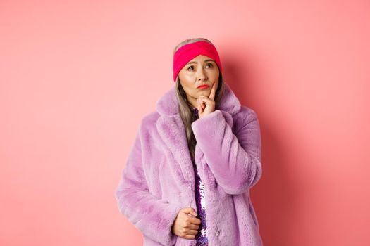 Funky old asian woman looking thoughtful and perplexed, standing in fake fur coat and thinking, making decision, standing on pink background