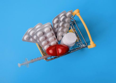 pills, vitamins and disposable syringe in shop trolley on blue background, the concept of heart treatment after vaccination, heart attack and other cardiac problems