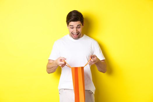Excited guy open bag with gift, looking inside with amazement and happy face, standing against yellow background