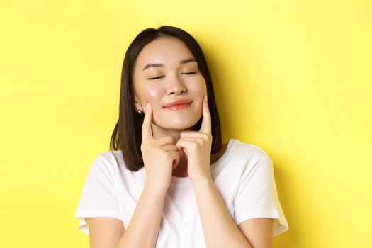 Beauty and skincare. Close up of young asian woman with short dark hair, healthy glowing skin, smiling and touching dimples on cheeks, standing over yellow background
