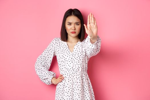 Angry asian woman tell stop, extend arm to prohibit or disapprove something, frowning displeased, standing over pink background