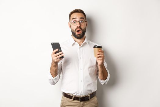 Image of handsome manage drinking coffee, reacting surprised to message on mobile phone, standing over white background