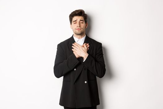 Portrait of dreamy and heartfelt bearded man in suit, close eyes and hold hands on heart, remember something, standing nostalgic over white background