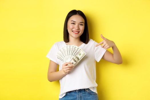 Young asian woman smiling, showing prize money, pointing finger at dollars, standing over yellow background