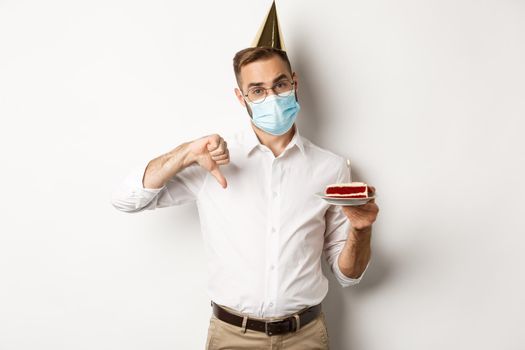 Coronavirus, quarantine and holidays. Man showing thumb down as disappointed with birthday party, wearing face mask and holding bday cake, white background