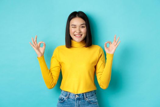 Cheerful asian female model showing okay gestures, smiling and looking impressed, praise product, standing over blue background