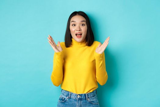 Image of happy asian woman clap hands and looking amazed, standing in yellow pullover over blue background