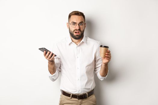 Image of man drinking coffee, feeling confused about strange message on mobile phone, standing over white background