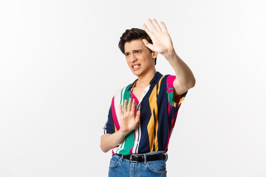 Timid and displeased androgynous man asking to stop, raising hands defensive and grimacing, standing over white background