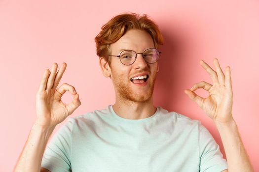 Cheeky redhaed guy in glasses assuring you, winking and showing okay signs, guarantee good quality, standing against pink background