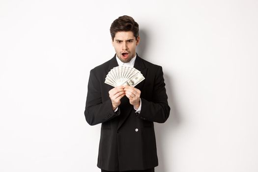 Portrait of handsome businessman in trendy suit, looking startled at money, won prize, standing over white background