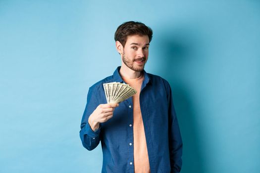 Unbothered rich guy showing cash and smiling, holding dollars in hand, standing on blue background