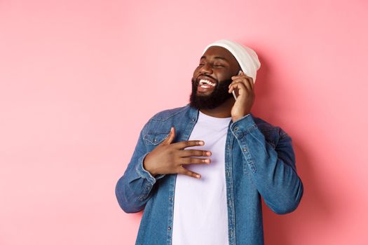 Happy african-american man talking on mobile phone, laughing and smiling, standing in beanie and denim shirt over pink background