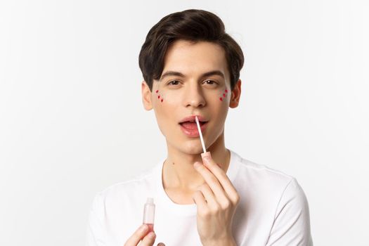 Close-up of gender fluid person with glitter on face, applying lip gloss lipstick and looking at camera, standing against white background