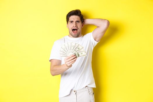 Frustrated man holding money, shouting and panicking, standing over yellow background