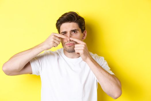 Young man squeezing pimple on nose, standing over yellow background. Concept of skin care and acne