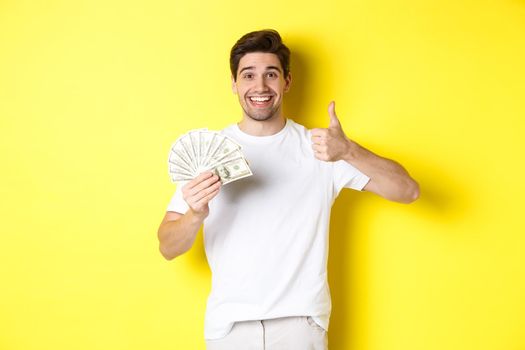 Satisfied buyer man showing thumb-up and holding money, shopping with cash, standing over yellow background