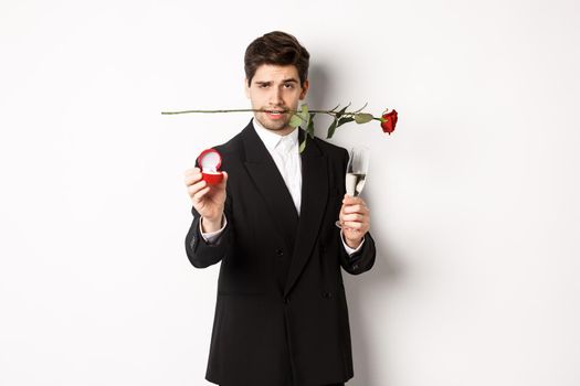 Passionate young man in suit making a proposal, holding rose in teeth and glass of champagne, showing engagement ring, asking to marry him, standing against white background