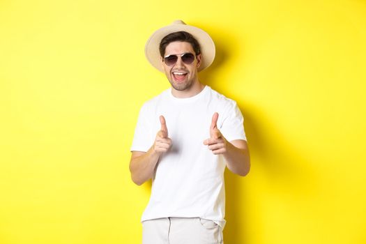 Confident and cheeky guy on vacation flirting with you, pointing finger at camera and winking, wearing summer hat with sunglasses, yellow background