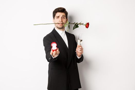 Romantic young man in suit making a proposal, holding rose in teeth and glass of champagne, showing engagement ring, asking to marry him, standing against white background