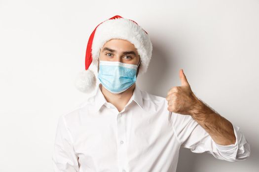 Concept of covid-19, social distancing and winter holidays. Satisfied man in face mask and santa hat showing thumb up, celebrating christmas with preventive measures, white background