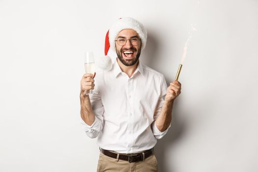 Winter holidays and celebration. Handsome bearded man having New Year party, holding firework sparkle and champagne, wearing santa hat, white background