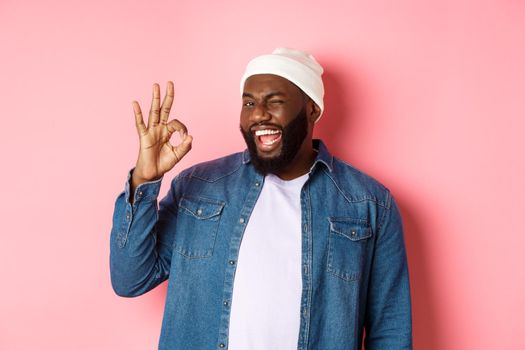 Happy smiling african american man showing okay sign, approve and praise good offer, standing over pink background
