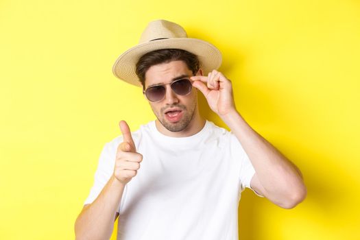 Concept of tourism and vacation. Cool and sassy man flirting with you, wearing sunglasses and pointing finger at camera, standing over yellow background